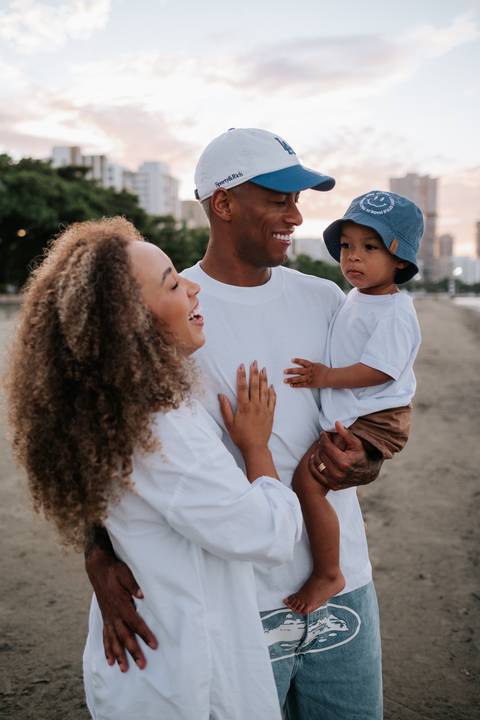 Foto aproximada do pai segurando o filho no colo e sorrindo para a mãe.'
