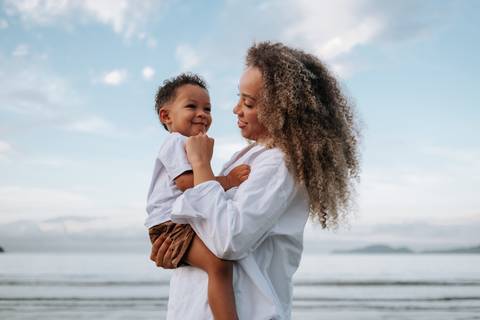 Foto da mãe segurando o filho no colo com o horizonte do mar ao fundo.'