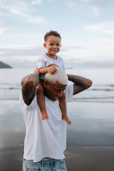 Foto do pai com o filho sentado em seus ombros na beira do mar.'