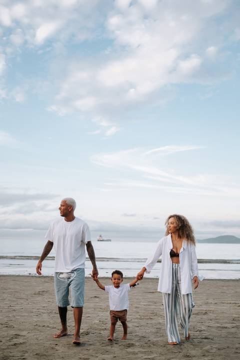 Foto panorâmica da família caminhando na praia sob um céu nublado e suave.'