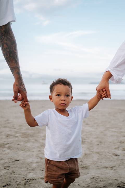 Foto de close do bebê segurando a mão do pai e da mãe na areia.'