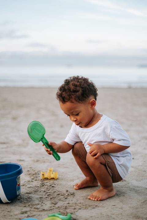 Foto aproximada do bebê agachado concentrado em brincar com a areia.'