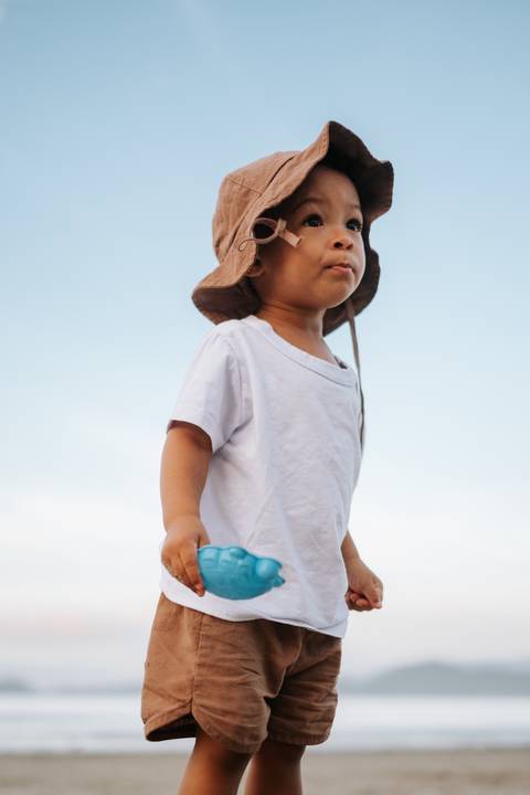 Foto do bebê de costas em pé na areia segurando seus brinquedos.'