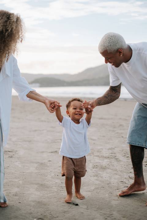 Foto do casal sorridente auxiliando os primeiros passos do bebê na praia.'