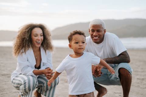 Foto oficial da família completa (pai, mãe e bebê) posando unida na praia.'