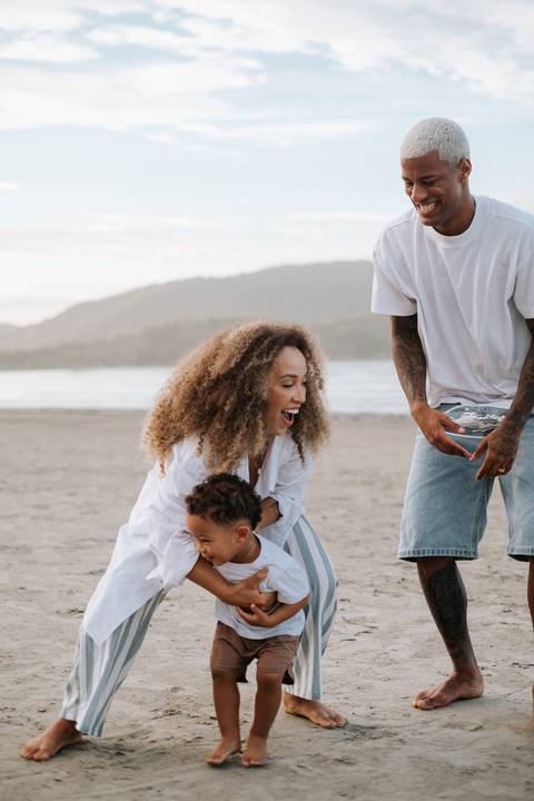 Foto da mãe agachada rindo enquanto o filho brinca na areia.'