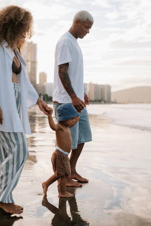 Foto em detalhe do bebê caminhando na areia molhada guiado pelos pais.'