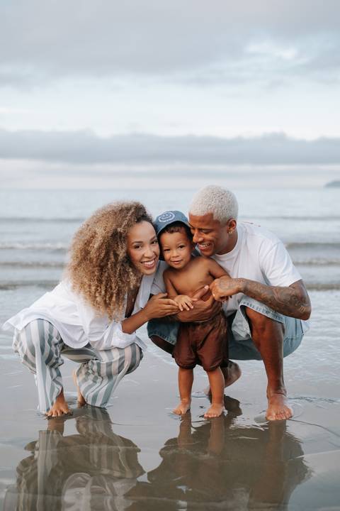 Foto do casal sorridente abraçado ao bebê na beira do mar.'