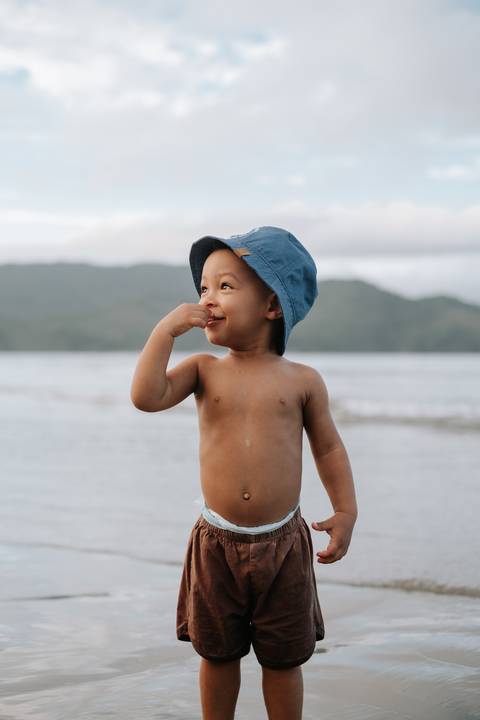 Foto do bebê fazendo uma pose fofa com a mão no rosto na beira-mar.'
