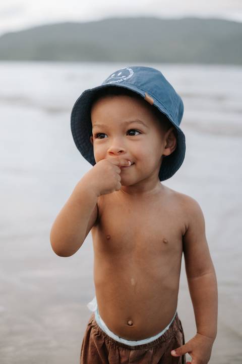 Foto de close do bebê com chapéu azul colocando a mão na boca.'