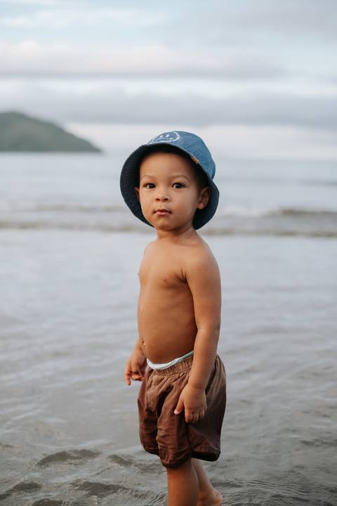 Foto do bebê em pé na areia molhada com o mar ao fundo.'