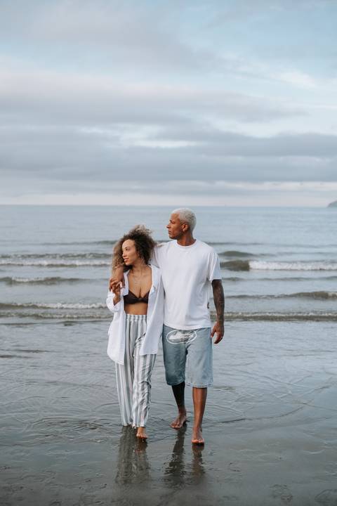 Foto do casal sorrindo e caminhando pela orla com céu nublado.'