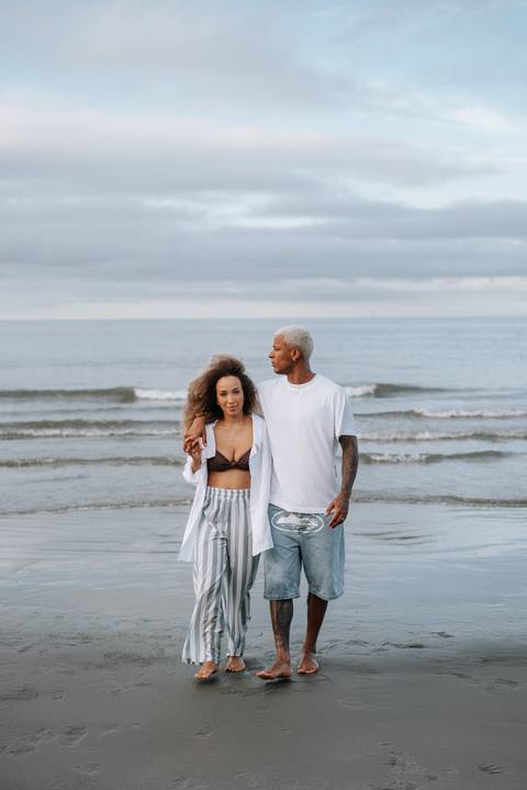 Foto aproximada do casal abraçado com o mar de cenário.'