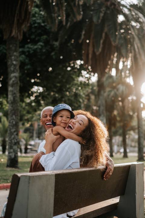 Foto do casal sorrindo com o filho de boné nos braços contra a luz do sol.'