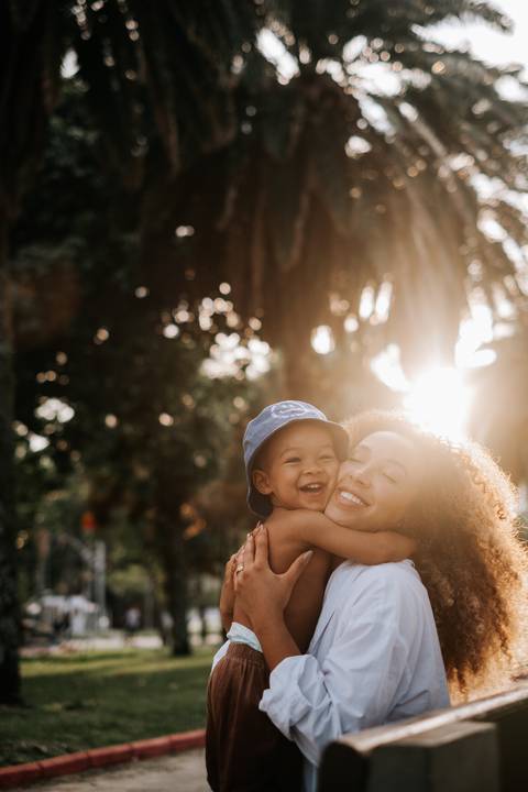 Foto aproximada da mãe e filho sorridente abraçada com forte luz solar de fundo.'