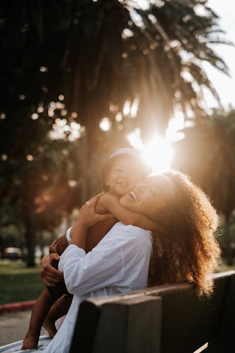 Foto em close da mãe e bebê com rostos colados e sorriso aberto sob o nascer do sol.'