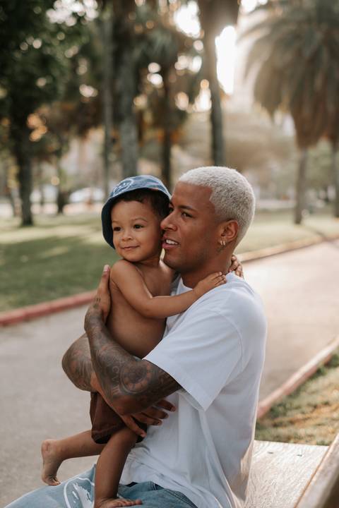 Foto de perfil do pai segurando o filho de boné no colo.'