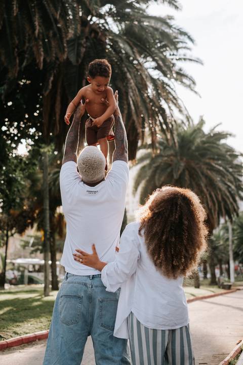 Foto da família caminhando unida pela orla arborizada; o pai carrega o filho.'