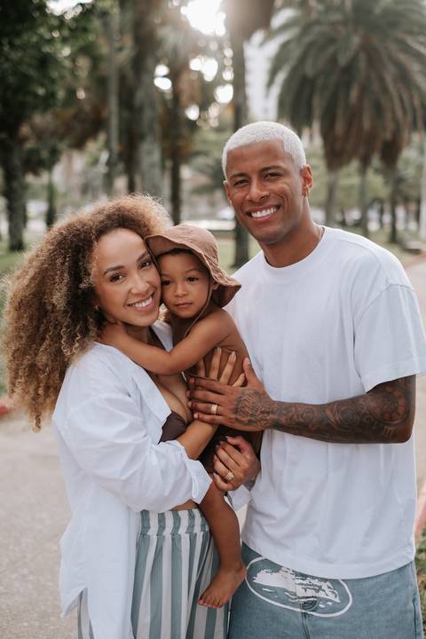 Foto oficial da família reunida (mãe, filho de boné e pai) posando sorridente na luz do sol.'