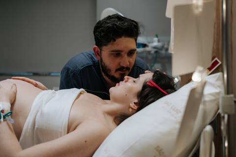 Foto do companheiro observando e apoiando a gestante deitada na cama de parto.'