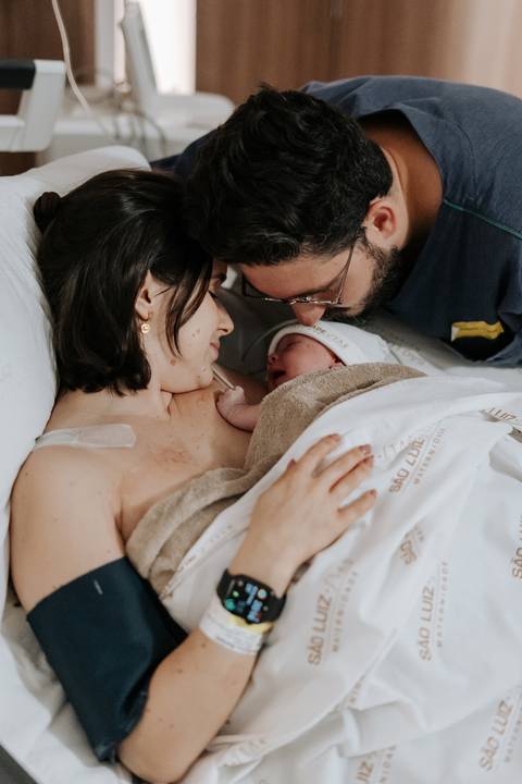 Foto do casal trocando olhares e sorrisos com o bebê nos braços.'