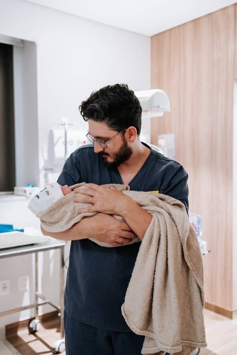 Foto do pai segurando o bebê no colo, envolto em uma manta, no hospital.'