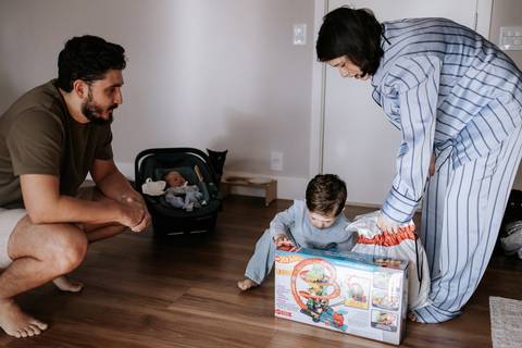 Foto da família reunida no chão da sala com o bebê no bebê-conforto e o irmão brincando com o novo presente. 
'