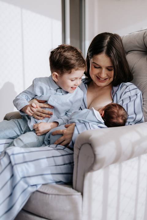 Foto da mãe amamentando o bebê enquanto abraça o filho mais velho no sofá.'