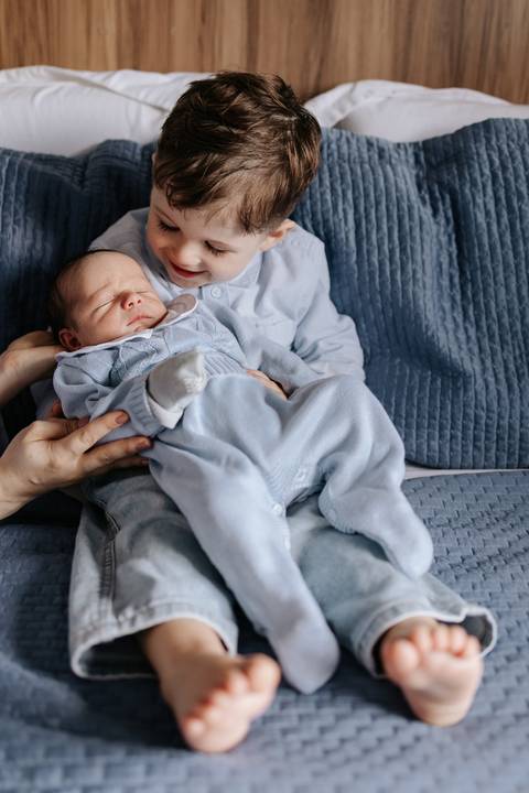 Foto do irmão mais velho segurando o bebê no colo com auxílio da mãe.'