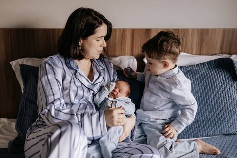 Foto da mãe sentada na cama com o recém-nascido e o filho mais velho.'