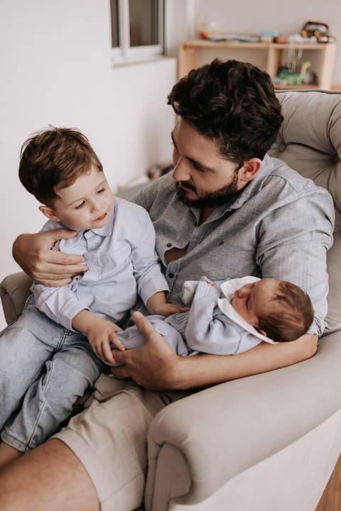 Foto do pai conversando com o primogênito com o recém-nascido no colo.'