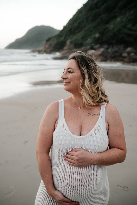 Foto da gestante sorridente olhando para o mar, vestindo vestido de crochê branco.'