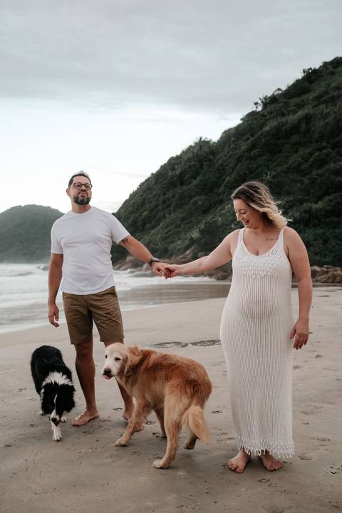 Foto do casal posando de frente na areia com seus cães ao redor.'