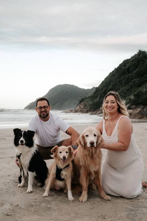 Foto da família completa: casal e seus quatro cães sentados na areia.'