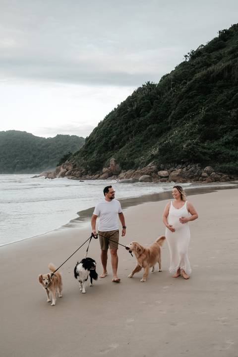 Foto do casal caminhando com os cães em uma paisagem de mar e montanhas.'