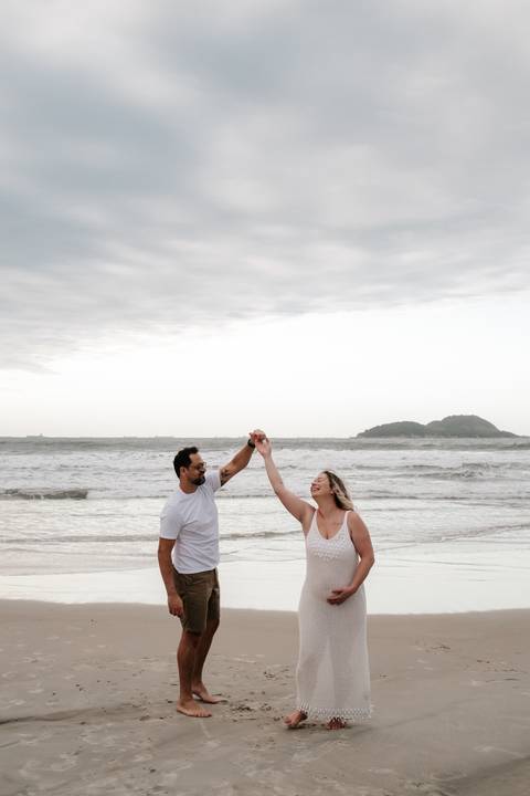 Foto panorâmica do casal dançando na beira do mar sob um céu nublado.'