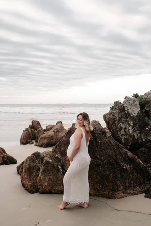 Foto da gestante de perfil posando em frente às pedras da praia.'