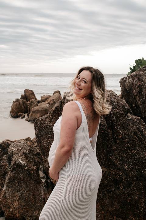 Foto da gestante olhando para trás e sorrindo, destacando o cenário rochoso.'