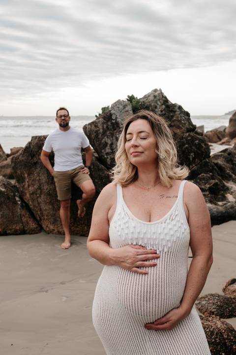 Foto da gestante com os olhos fechados sentada nas pedras, com o parceiro ao fundo.'