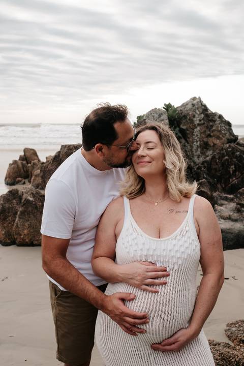 Foto aproximada do parceiro beijando o rosto da gestante em frente às rochas.'