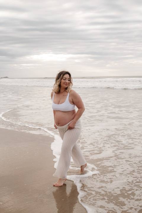 Foto da gestante caminhando na areia vestindo top branco e calça de rede.'