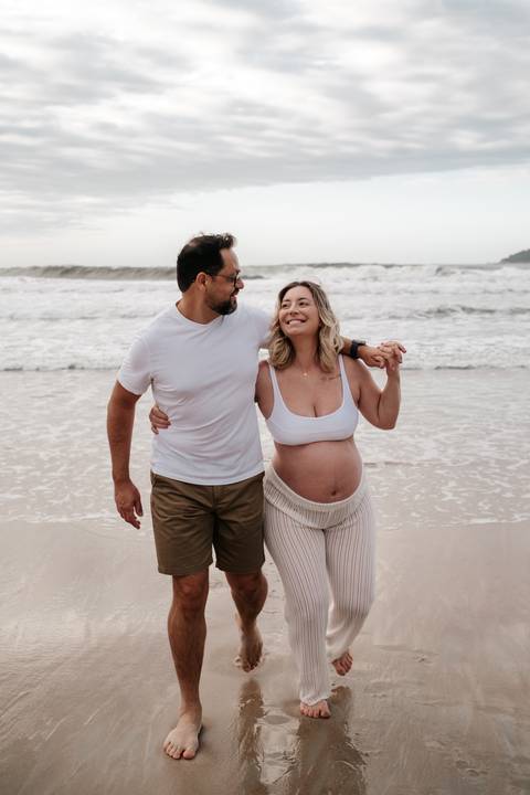 Foto do casal caminhando de braços dados na beira da água, sorrindo.'