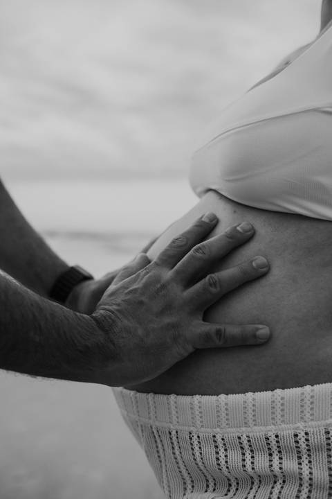 Foto em preto e branco focado no detalhe das mãos sobre a barriga da gestante.'