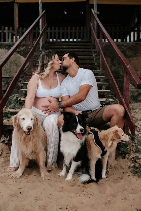 Foto do casal sentado na escada de madeira da praia com seus três cães.'