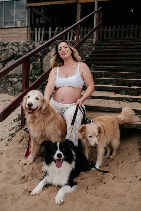 Foto da gestante em pé exibindo a barriga rodeada pelos pets da família.'