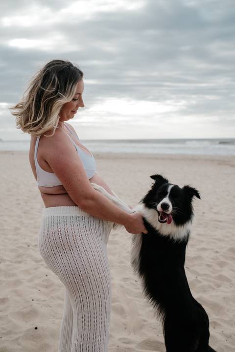 Foto de perfil da gestante sorrindo para seu cão Border Collie na areia.'