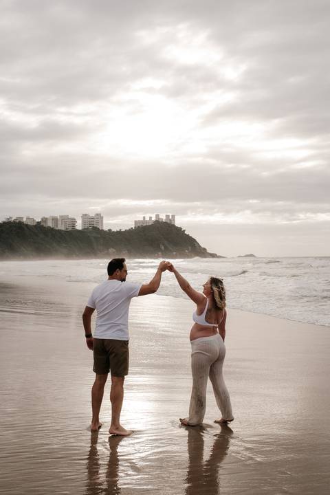 Foto panorâmica do casal de mãos dadas fazendo um arco com os braços na orla.'