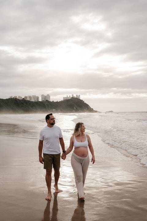 Foto do casal caminhando na beira do mar sob o céu nublado do entardecer.'