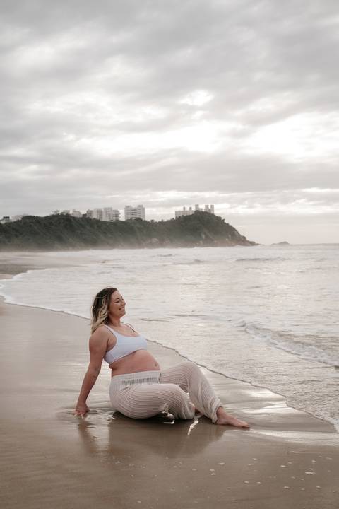 Foto da gestante sorridente sentada na areia com as ondas ao fundo.'