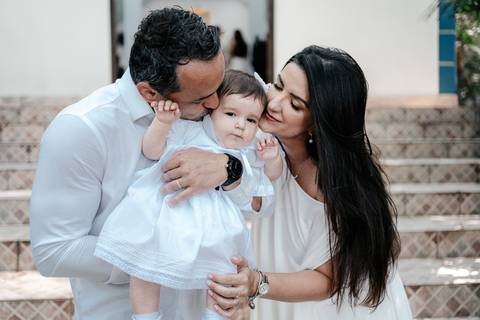 Retrato do casal beijando a bebê vestido com roupa de batizado branca nos degraus da capela.'
